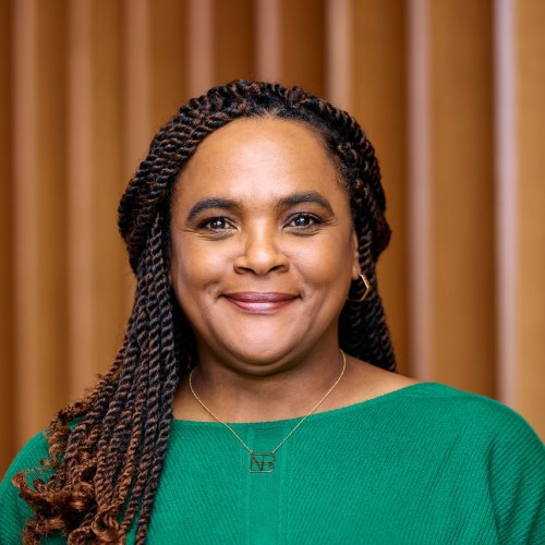 Portrait of Brenda Ice, smiling, wearing a green top, standing in front of a wood-paneled background.
