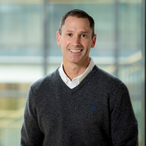 A smiling man wearing a dark sweater over a white collared shirt, standing in front of a softly blurred glass wall background.
