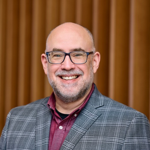 A smiling man with glasses wearing a gray plaid blazer over a burgundy shirt, standing in front of a brown vertical-striped background.