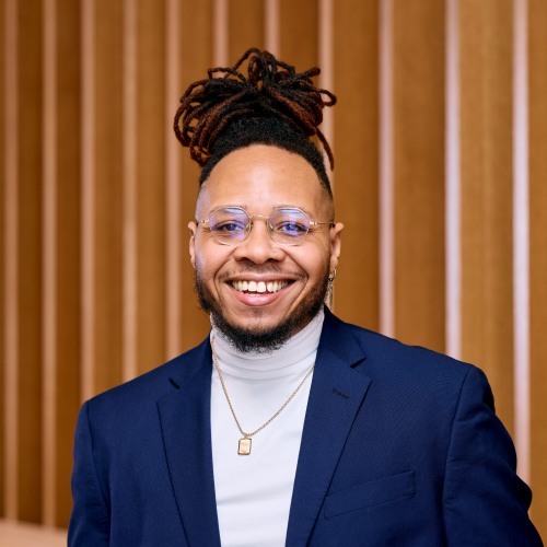 A smiling person with glasses and styled dreadlocks in a high bun, wearing a blue blazer over a light turtleneck sweater, standing in front of a brown vertical-striped background.