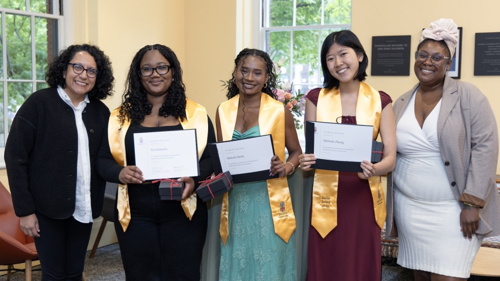 Three Joslin award recipients, two with dark, curly hair and the third with dark, short hair, stand in the center of a group holding their awards and each wearing a gold "Joslin Award" stole, surrounded by the director and assistant director of the Sarah Doyle Center for Women and Gender.