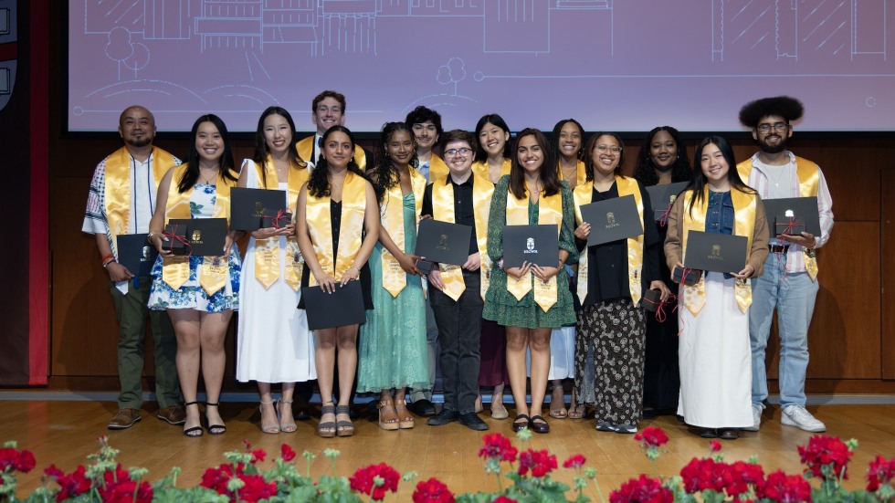 The 2025 Campus Life Award winners are shown standing in a line, wearing their bright yellow Joslin recipient stoles, before a red backdrop featuring a white outline of the Brown campus skyline.